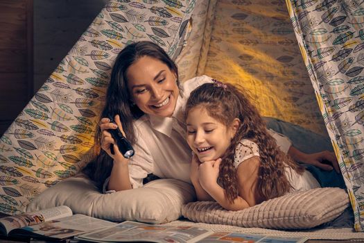 Mother and daughter are sitting in a teepee tent, reading stories with the flashlight and laughing. Happy family.