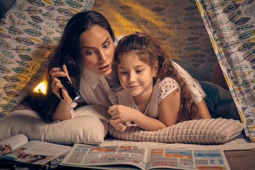 Mother and her pretty daughter are sitting in a teepee tent, reading stories with the flashlight. Happy family.