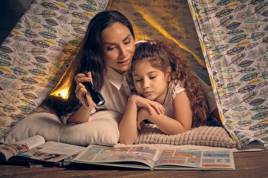 Mother and her charming daughter are sitting in a teepee tent, reading stories with the flashlight. Happy family.