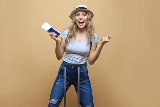 Cheerful blonde woman in summer clothes posing with baggage and holding passport with tickets over beige background