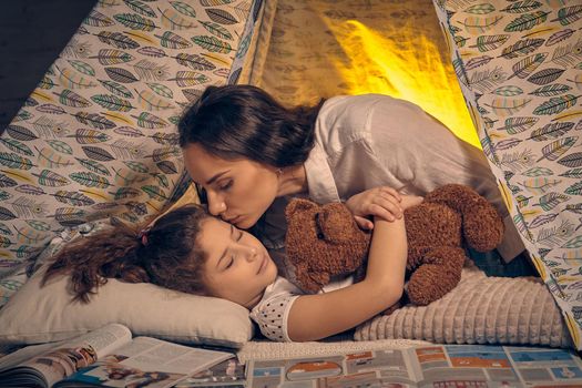 Young mother and her pretty daughter are in a teepee tent with some pillows. Girl is sleeping and hugging a toy bear. Mom is kissing her. Happy family.