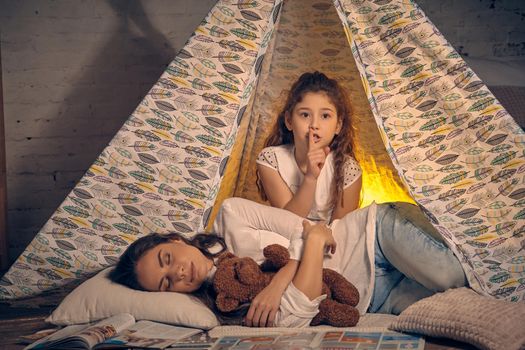 Young mother and her pretty daughter are in a teepee tent with some pillows. Mom is sleeping with a toy bear. Daughter is showing sign quieter. Happy family.