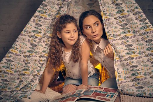 Young mother and her little daughter are sitting in a teepee tent with some pillows and looking away. Happy family.