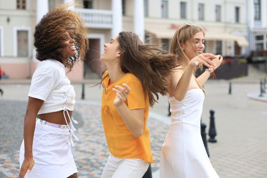 Three young smiling hipster women in summer clothes posing on street.Female showing positive face emotions. Dancing