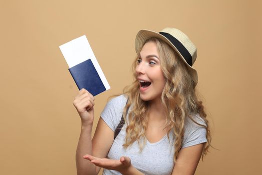 Beautiful blonde woman wearing summer clothes posing with passport with tickets over beige background