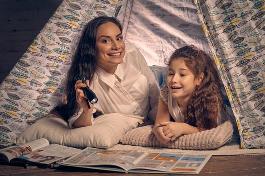 Mother and daughter are sitting in a teepee tent, reading stories with the flashlight and smiling. Happy family.