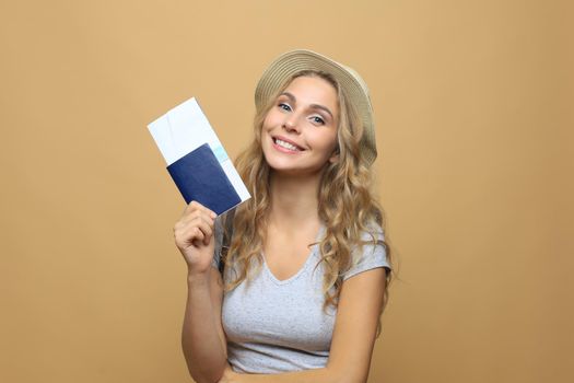 Beautiful blonde woman wearing summer clothes posing with passport with tickets over beige background