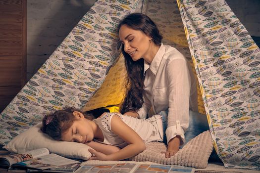 Mother and her daughter are in a teepee tent with some pillows. Mom is looking at her daughter while she is sleeping. Happy family.