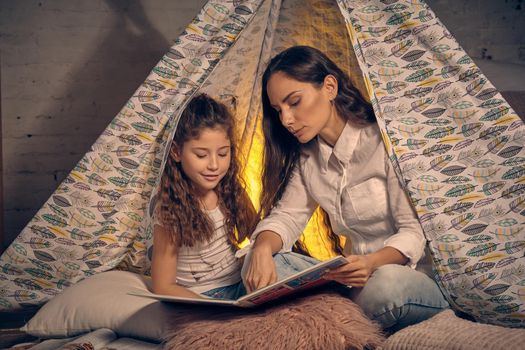 Mother and daughter are sitting in a teepee tent with some pillows and reading stories. Mom is showing something in the book. Happy family.