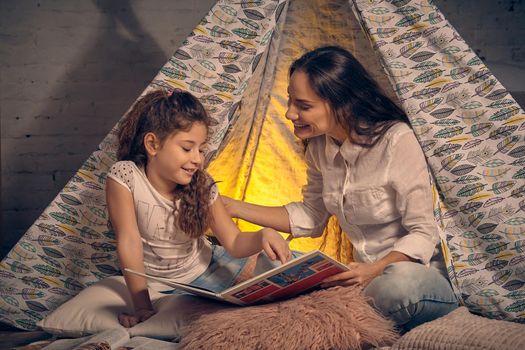 Mother and daughter are sitting in a teepee tent with some pillows, reading stories and smiling. Happy family.