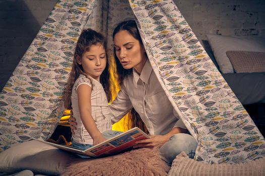 Mother and daughter are sitting in a teepee tent with some pillows and reading stories. Mom is hugging her princess. Happy family.