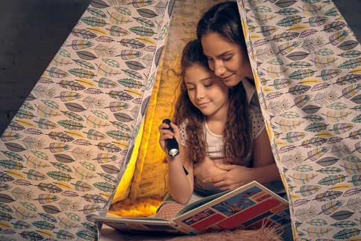 Young mother and her beautiful daughter are sitting in a teepee tent, reading stories with the flashlight. Happy family.