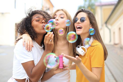 Three happy beautiful women blowing soap bubbles in city street