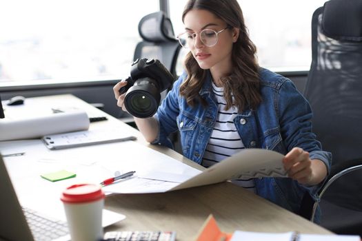 Young architect holding digital camera and looking on blueprint while working in the creative office