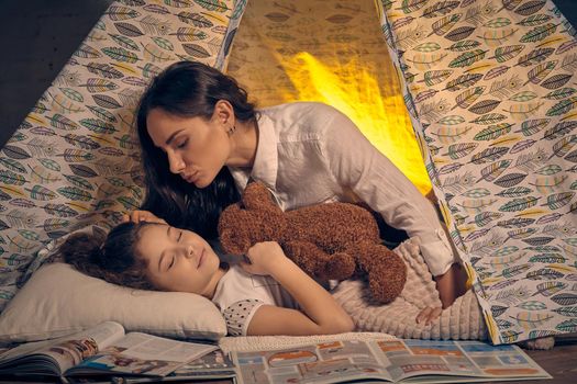 Young mother and her pretty daughter are in a teepee tent with some pillows. Girl is sleeping and hugging a toy bear. Mom is looking and stroking her. Happy family.