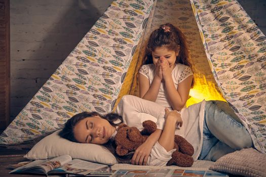 Young mother and her pretty daughter are in a teepee tent with some pillows. Mom is sleeping with a toy bear. Daughter is smiling and looking at her. Happy family.