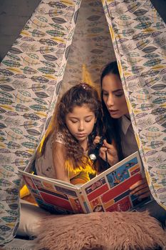 Mother and daughter are sitting in a teepee tent with some pillows, reading stories with the flashlight. Happy family.