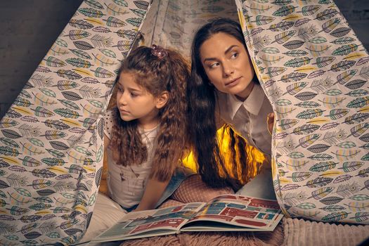 Young mother and her daughter are sitting in a teepee tent with some pillows and looking around. Happy family.