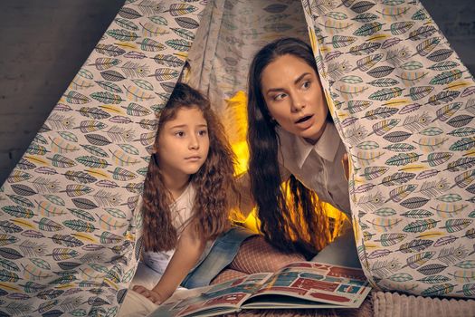 Mother and her daughter are sitting in a teepee tent with some pillows and looking wondered by something. Happy family.