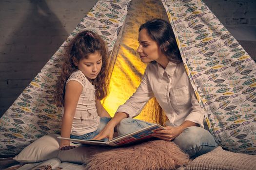 Mother and daughter are sitting in a teepee tent with some pillows and reading stories. Mom is showing something in the book and looking at her. Happy family.