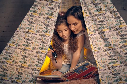 Young mother and daughter are sitting in a teepee tent with some pillows, reading stories with the flashlight. Happy family.