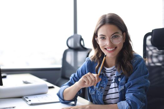 Beautiful young woman looking at camera and holding pencil while sitting in modern office