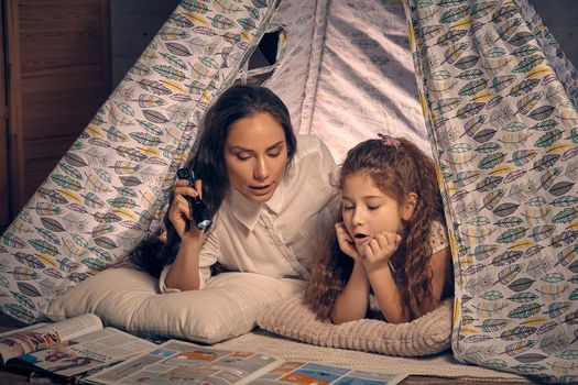 Mother and daughter are sitting in a teepee tent, reading stories with the flashlight. Happy family. They are reading together.