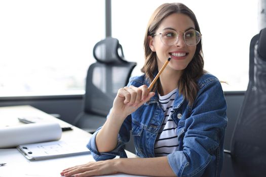 Beautiful young woman looking at camera and holding pencil while sitting in modern office