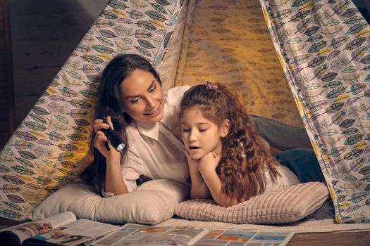 Mother and daughter are sitting in a teepee tent, reading stories with the flashlight. Happy family.