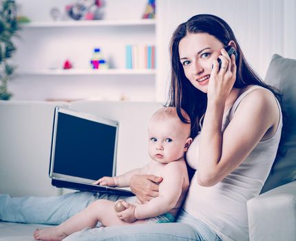 mother holding one year old baby in his arms,talking on his cell phone and working on laptop sitting on sofa in living room