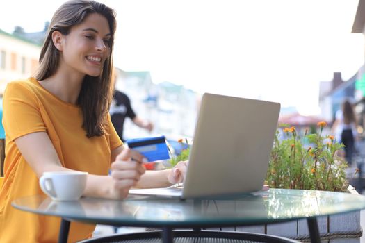 Happy natural brunette using credit card and laptop in cafe outdoors