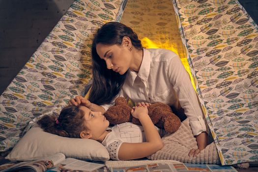 Young mother and her daughter are in a teepee tent with some pillows. Girl is lying and hugging a toy bear. They are looking at each other and mom is stroking her. Happy family.