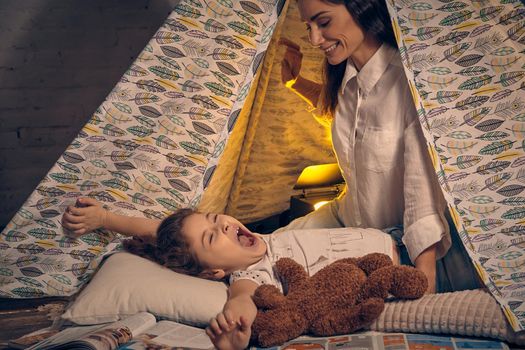 Young mother and her pretty daughter are in a teepee tent with some pillows. Girl is lying with a toy bear and yawning. Mom is looking at her. Happy family.