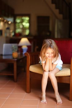 Female blonde kid sitting in wooden chair in living room. Concept of child model and cozy interior.