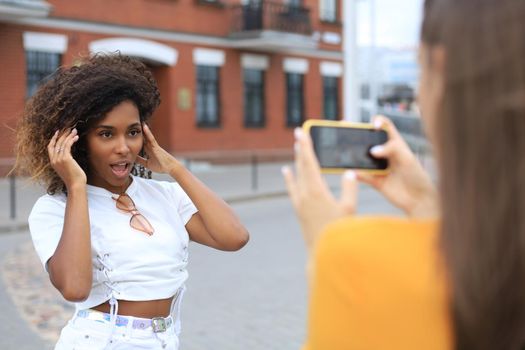 Two young smiling hipster women in summer clothes posing on street.Female showing positive face emotions