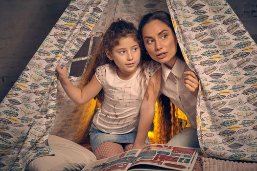 Young mother and her pretty daughter are sitting in a teepee tent with some pillows and looking away. Happy family.