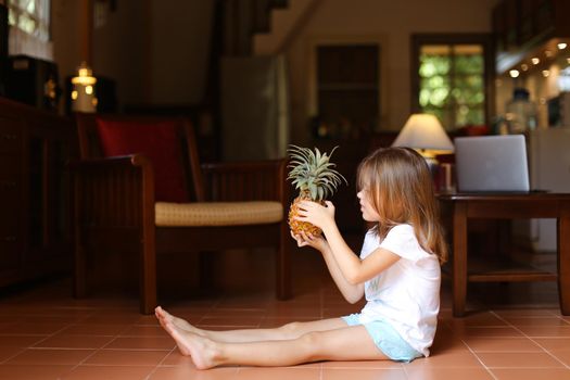 Little female american kid sitting on floor in living room and playing with pineapple, laptop in background. Concept of health life, fruit and childhood.