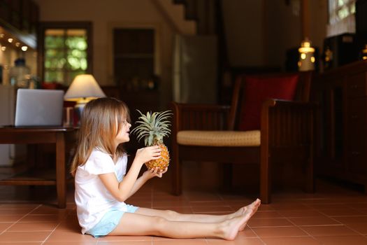 Little european female kid sitting on floor in living room and playing with pineapple, laptop in background. Concept of health life, fruit and childhood.