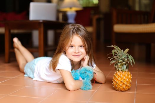 Little smiling girl lying on floor and playing with pineapple and toy, laptop in background. Concept of health vegeterian life and childhood.