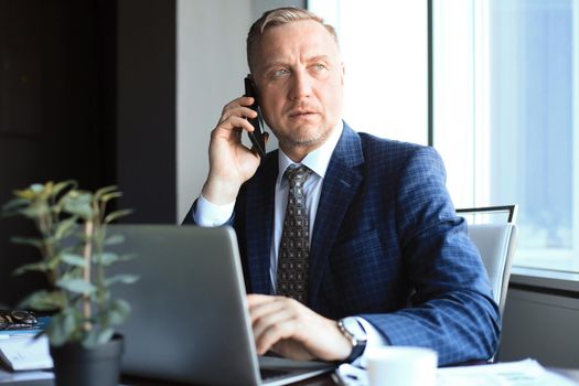 Middle aged businessman with laptop talking on telephone in modern office
