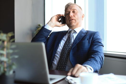 Middle aged businessman with laptop talking on telephone in modern office