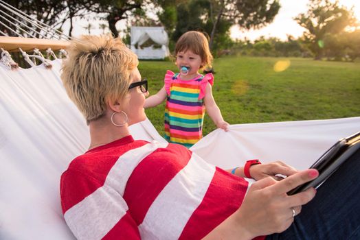 Happy mother and her little daughter enjoying free time using tablet computer while relaxing in a hammock during sunny day on holiday home garden