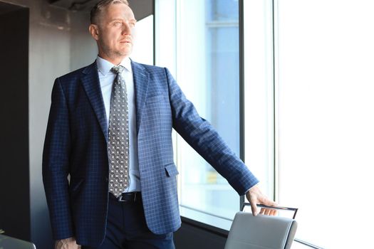 Mature businessman in a corporate suit standing in office and looking away through large windows optimistically