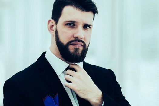 portrait of a successful businessman adjusting his necktie while standing near a window in bright office.the photo has a empty space for your text