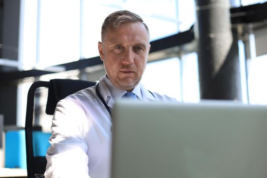 Senior physician in white robe with stethoscope using laptop