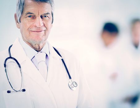 Portrait of an experienced physician in age, in background work team. He is smiling and looking into the camera