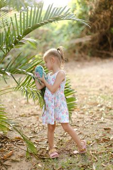 Little girl playing with toy and standing barefoot near palm leaves. Concept of vacations with children in exotic resorts, tour to Thailand with kids.