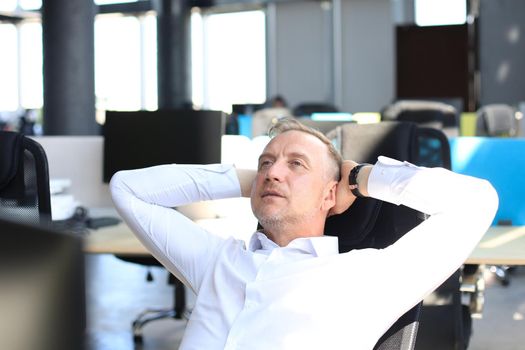 Middle aged modern businessman keeping hands behind head and smiling while sitting in the office