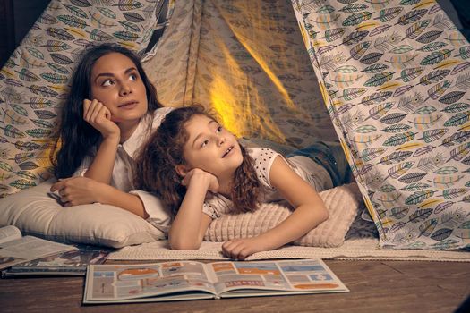 Mother and daughter are sitting in a teepee tent with some pillows and reading stories. They both are looking up. Happy family.