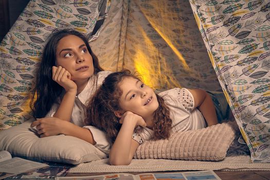 Young mother and daughter are sitting in a teepee tent with some pillows and reading stories. They both are looking up. Happy family.